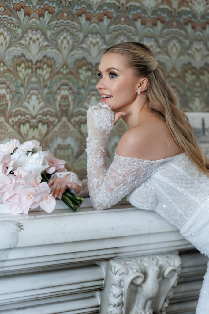 Braut im bestickten Spitzenkleid mit Brautstrauß in Blush am historischen Kamin der Villa Aurea in Potsdam – Komplettplanung durch Spreejuwel Hochzeiten
