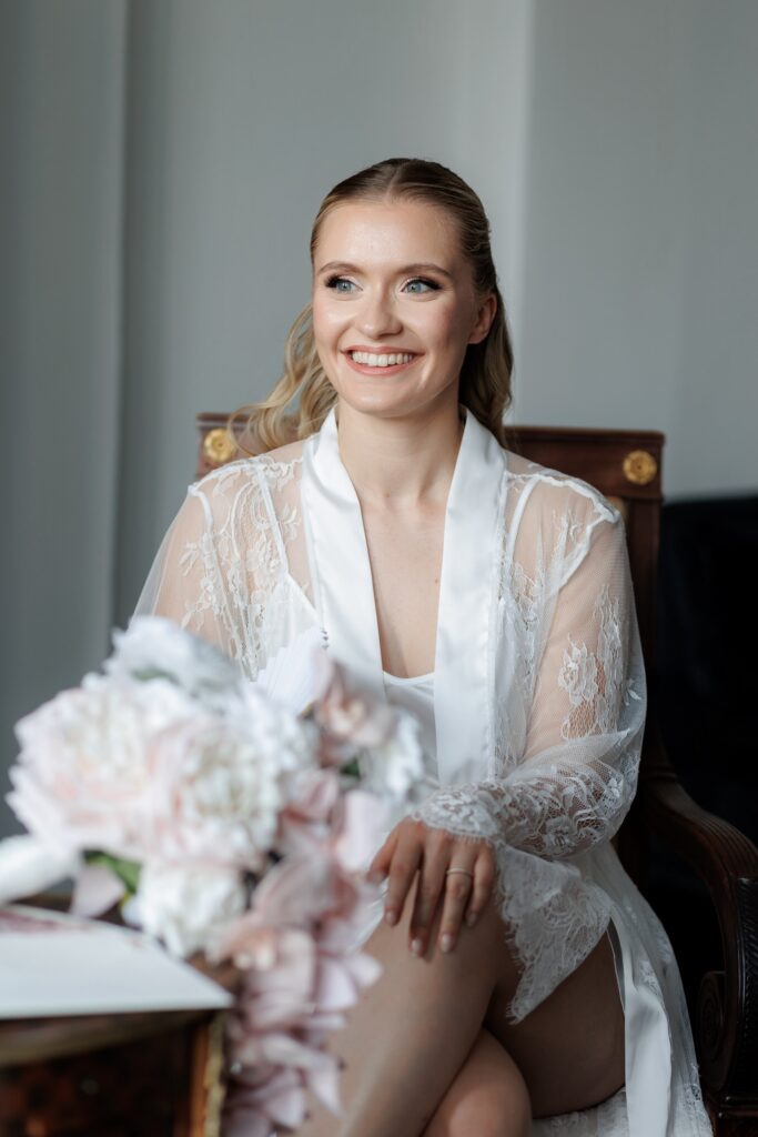 Braut im Spitzenmorgenmantel beim Getting Ready am Fenster, im Vordergrund Brautstrauß in Blush – Tagesbegleitung durch Spreejuwel Hochzeiten