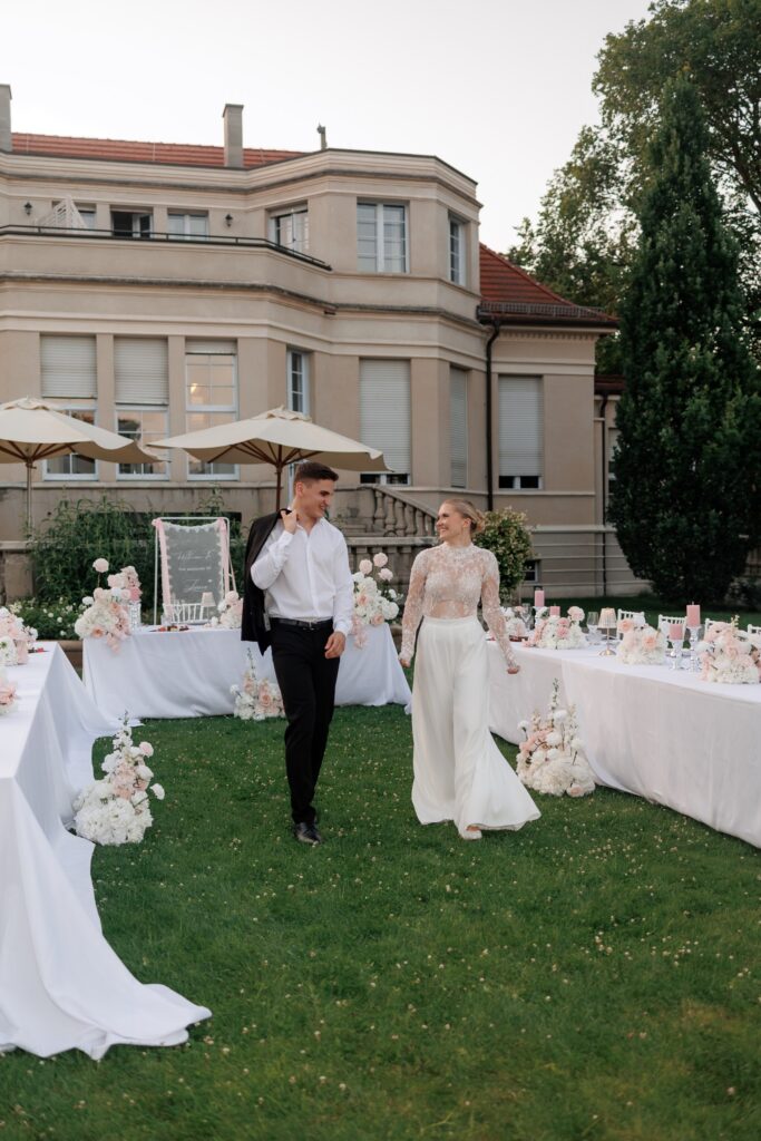 Brautpaar geht über den Rasen an der festlich gedeckten Tafel vorbei, im Hintergrund die Villa Aurea in Potsdam – Komplettplanung durch Spreejuwel Hochzeiten